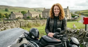 A candid lifestyle shot of a female motorcyclist in an RST leather jacket standing by her Triumph bike on a country road in the Yorkshire Dales, featuring a classic red post box.