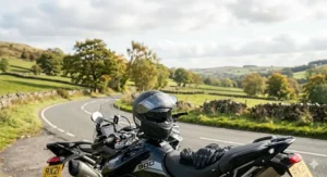 A SHARP 5 star helmet resting on a motorcycle seat alongside leather gloves, set against a British landscape with a yellow rear registration plate visible.