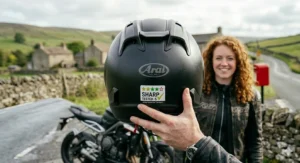 A detailed close-up of a 5-star SHARP tested safety rating sticker on the rear of a matte black full face crash helmet, with a Triumph motorcycle and stone cottages blurred in the background.