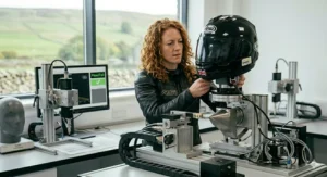 A female safety engineer in an RST jacket meticulously fitting a black Arai helmet to a linear actuator testing machine for a SHARP impact assessment.