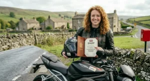 A British rider presenting an official SHARP certificate of excellence next to her 5-star rated full face helmet and motorcycle in a rural UK setting.