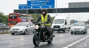 A British commuter riding a maxi-scooter on a damp UK dual carriageway wearing a quiet bluetooth motorcycle helmet.