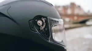 An exploded view of the left-side quick-release visor mechanism on the matte black polycarbonate helmet, with the clear visor detached.