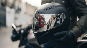 A detailed close-up shot of a matte black motorcycle helmet with a highly reflective silver mirror visor, held by a gloved hand. The visor clearly reflects a miniature London scene, including a red double-decker bus and a black taxi. The texture of the helmet and glove is sharp under natural light.