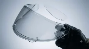A detailed close-up studio photograph of the interior surface of the silver mirror motorcycle helmet visor. The gloved hand holds the visor, revealing the small clear Pinlock pins along the edge. A transparent Pinlock anti-fog insert is shown partially aligned, ready for installation.