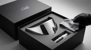A detailed close-up studio photograph of an opened premium matte black presentation box. Inside, the silver mirror motorcycle helmet visor is securely nestled in custom-cut foam. Beside it, a folded microfibre cleaning cloth and a small sealed sachet of anti-fog spray are presented. The rider's gloved hand is lifting the cloth.